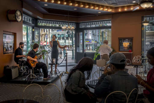 Live music at Cafe Beignet Sheraton. A singer, drummer, and guitarist perform onstage in front of open doors to the street.