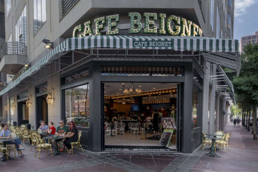 The exterior of Cafe Beignet Sheraton. Guests eat and chat on tables outdoors and inside the cafe.
