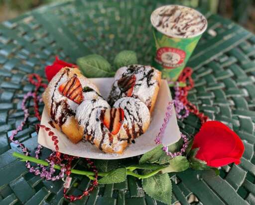 Beignets topped with strawberries and chocolate drizzle at Cafe Beignet, with a coffee, a rose, and heart-shaped beads arranged around them.