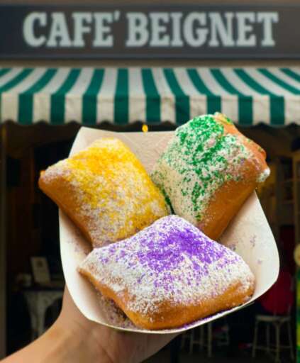 King Cake Beignets with purple, green, and gold sprinkles on top, in front of the Cafe Beignet sign.